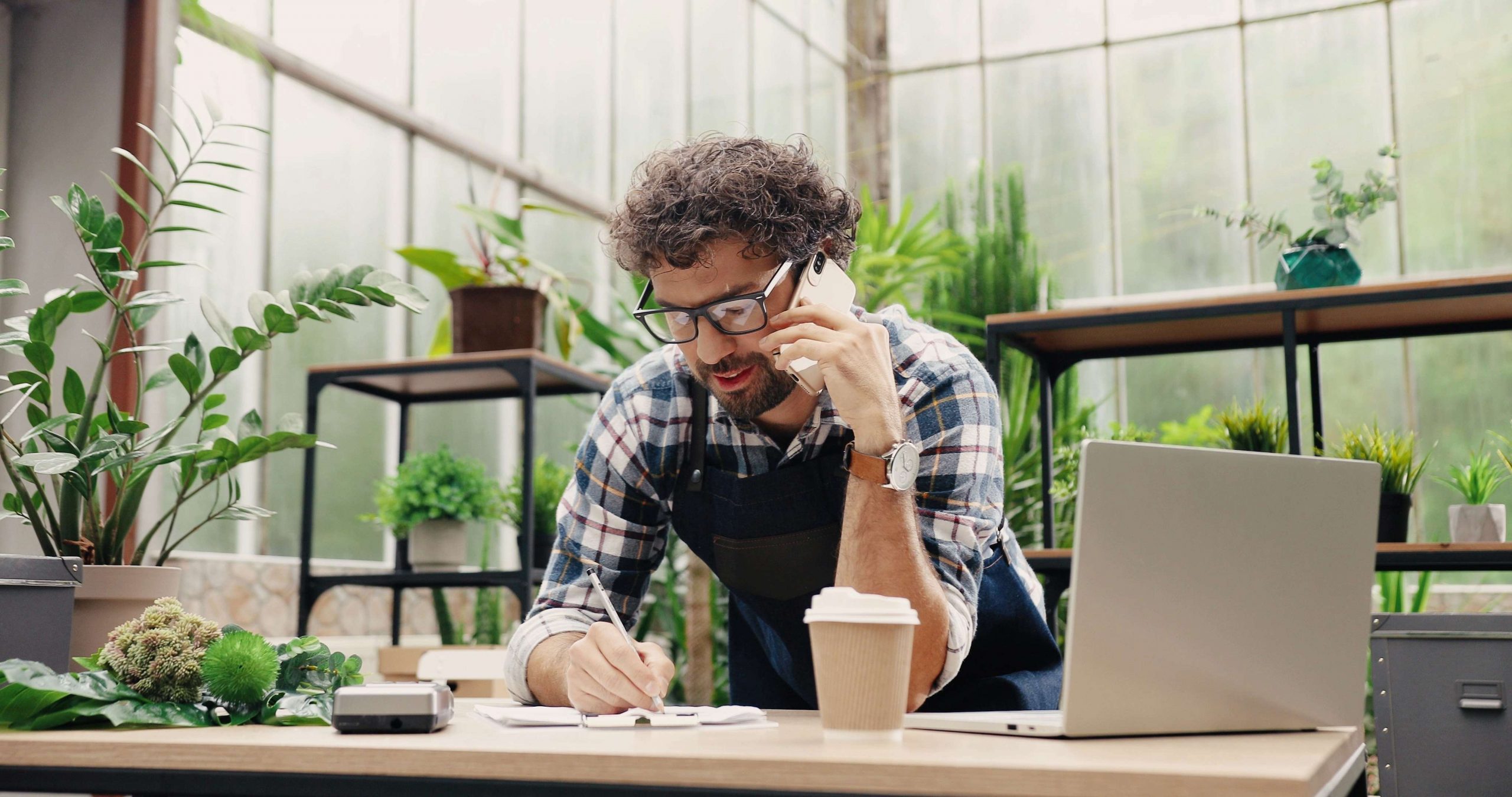 Florist talking on the phone while standing in apron in small floral centre and writing down order details