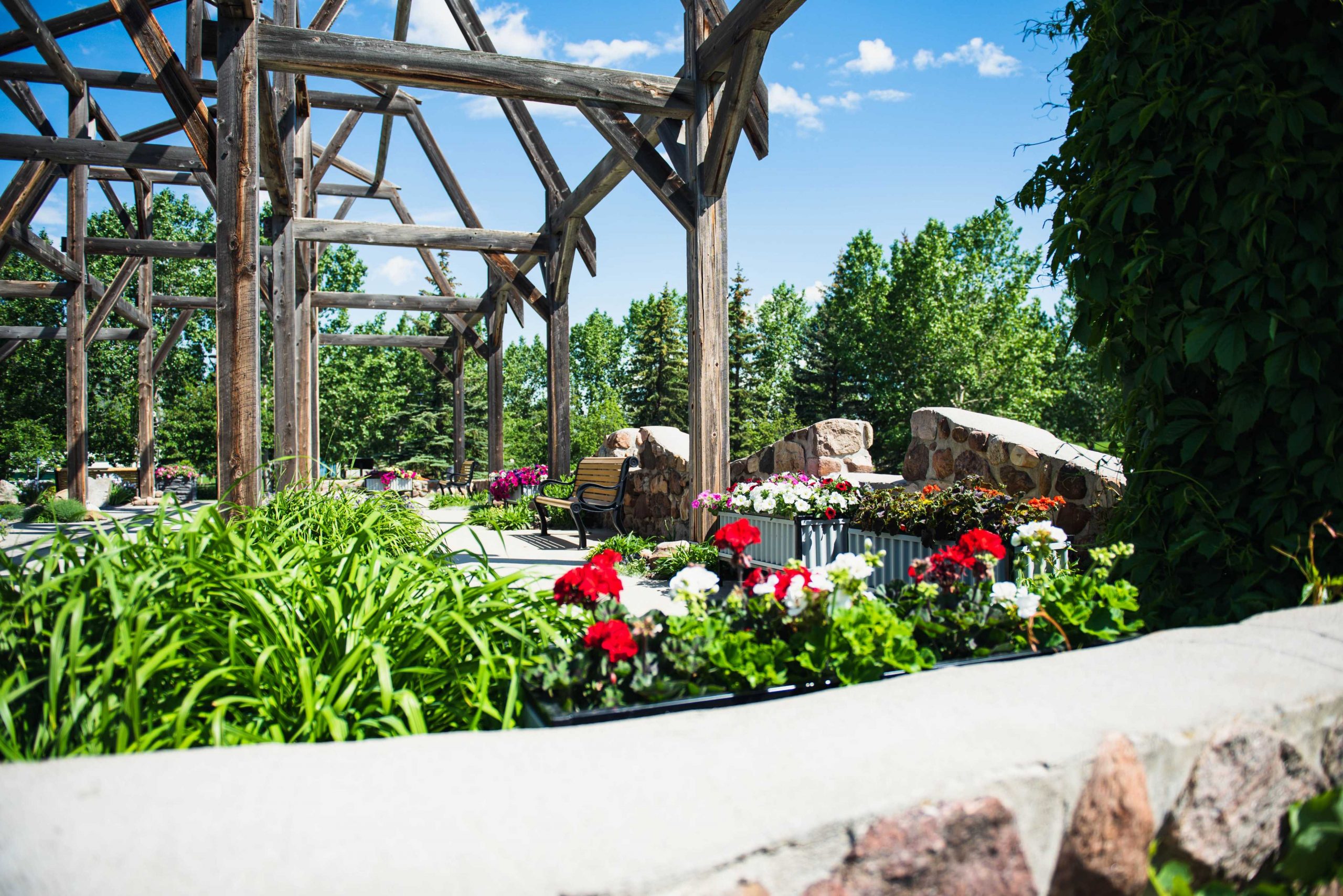 Wooden pavilion structure surrounded by colorful flowers and greenery under a clear blue sky.
