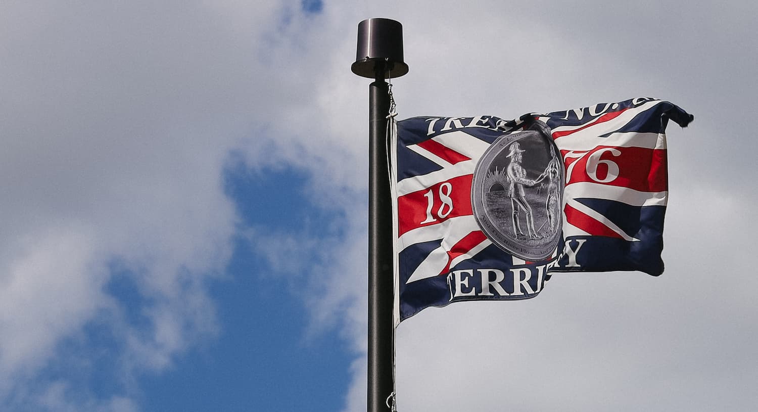 A flag with the design of a Union Jack and the text "1865" against a partly cloudy sky.